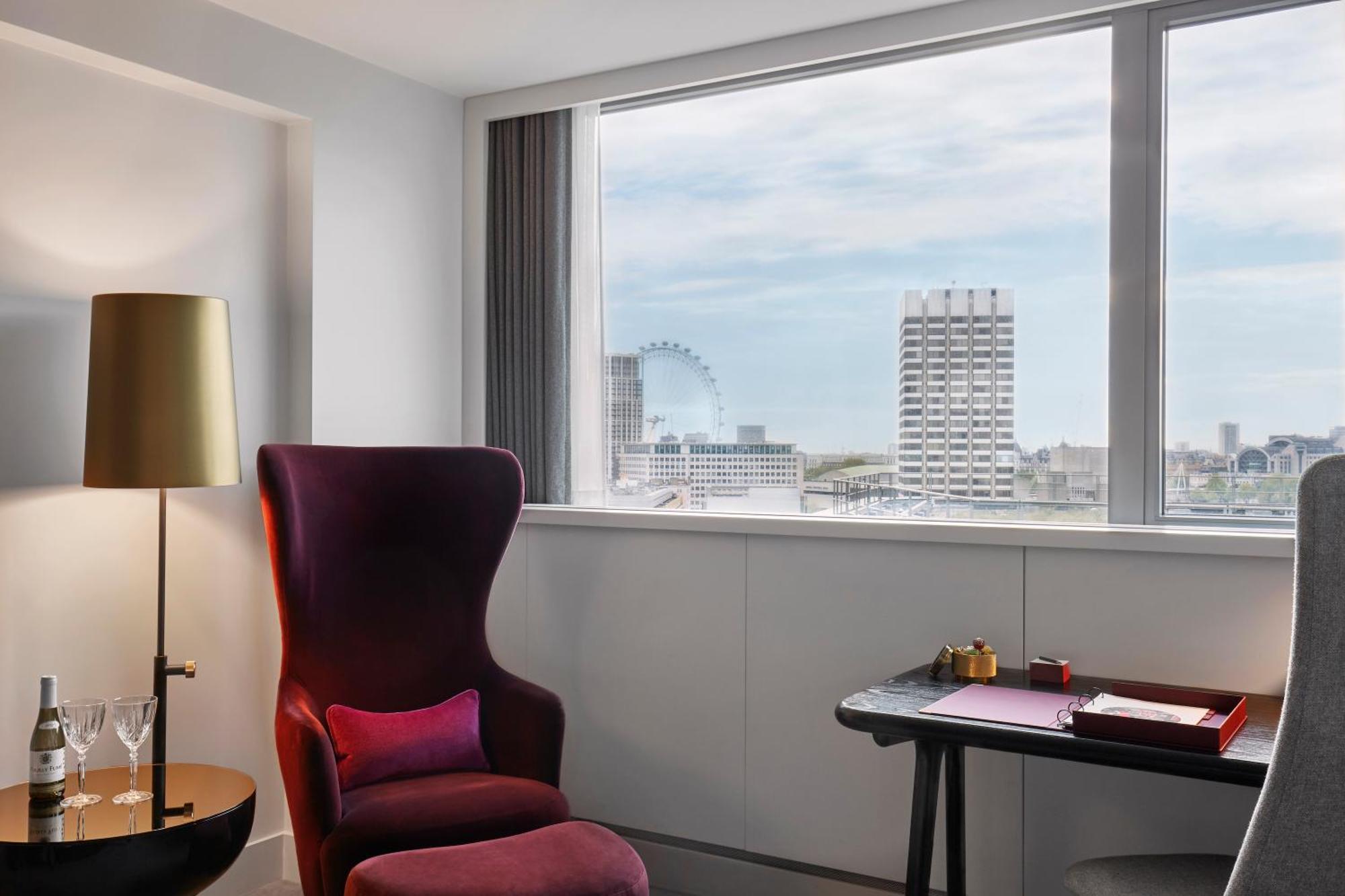 Sea Containers London Hotell Eksteriør bilde The photo shows a modern and stylish hotel room featuring a large window with a view of the city skyline. There is a plush, deep purple armchair with a pink cushion in one corner, creating a cozy seating area. Next to the chair, there's a sleek floor