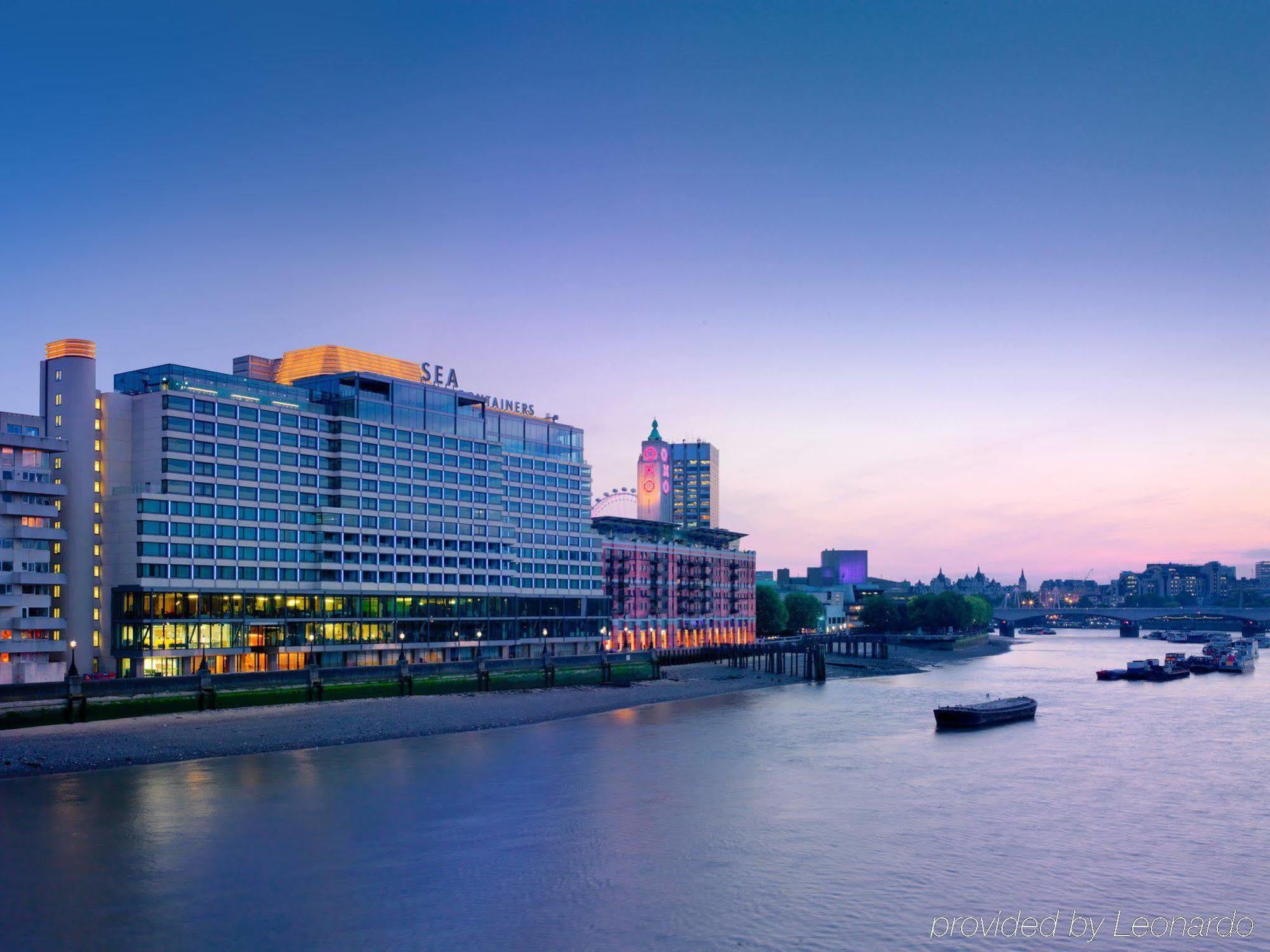 Sea Containers London Hotell Eksteriør bilde The photo depicts a waterfront scene featuring a modern hotel building next to a river. The hotel has a sleek, contemporary design, with multiple stories and large windows reflecting the evening light. The sky transitions from a soft blue to warmer h